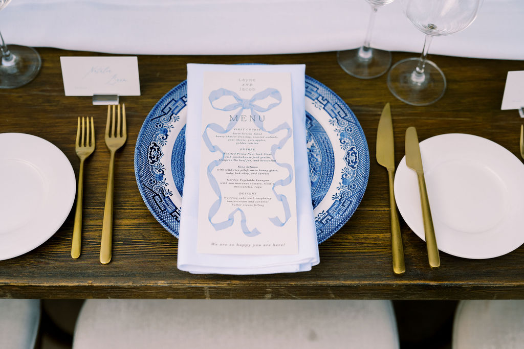 A formal table setting with a blue-patterned plate, gold utensils, a white napkin, a printed menu, wine glasses, and name cards on a wooden table.