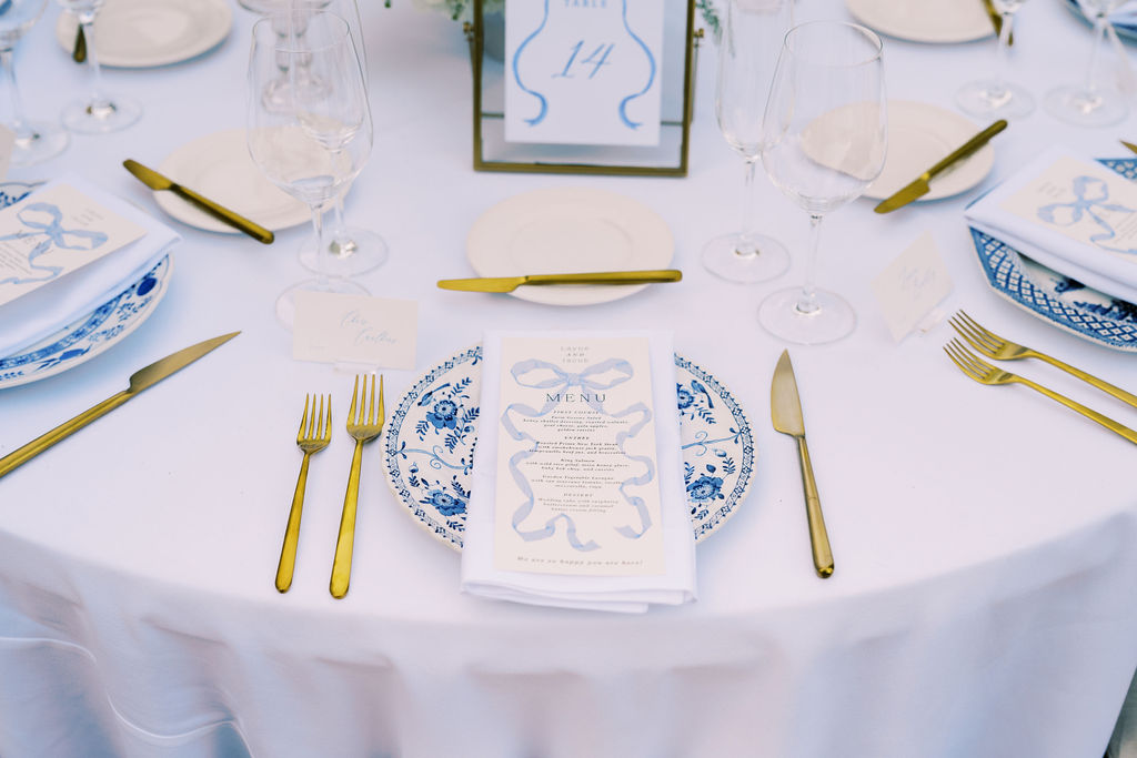 A formal table setting with blue and white plates, gold utensils, wine glasses, and a printed menu on a white tablecloth.
