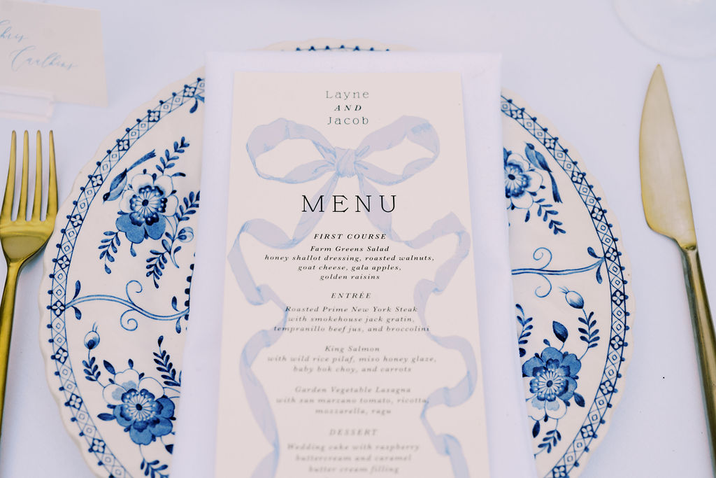 A printed wedding menu with blue ribbon design is placed on a white plate with blue floral patterns, accompanied by gold cutlery.