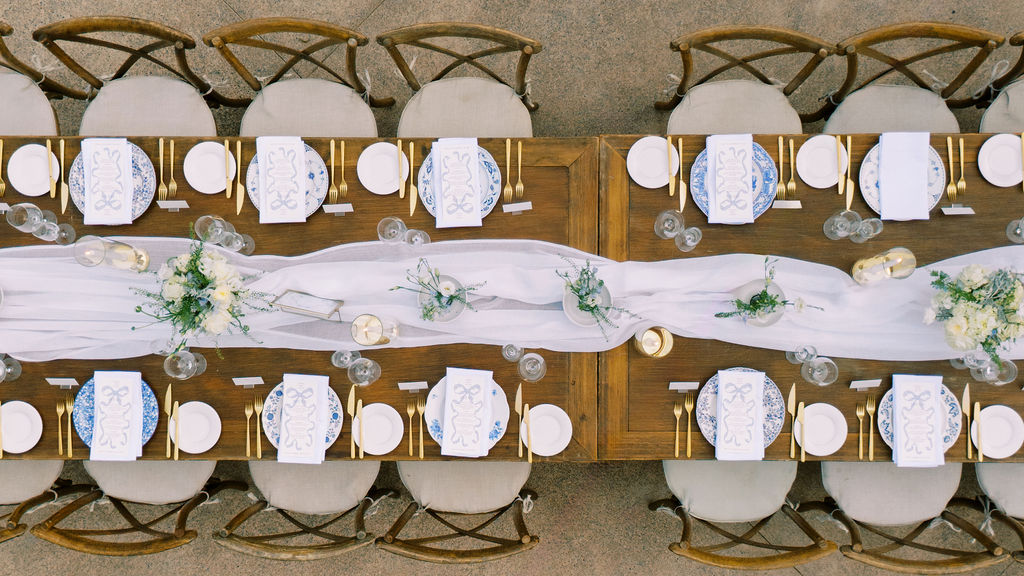 Aerial view of a long wooden dining table set for an event with white plates, blue napkins, gold cutlery, floral centerpieces, and a white table runner.
