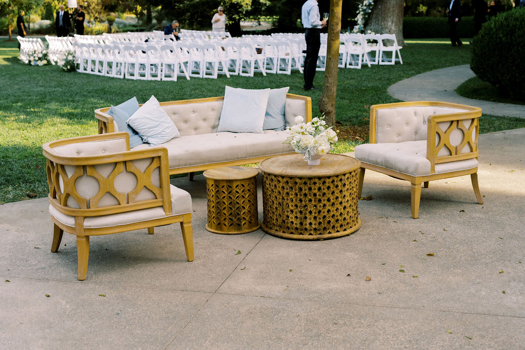 Outdoor seating area with wooden chairs, a loveseat, a round table, and white cushions; white chairs arranged in rows are visible in the background on grass.
