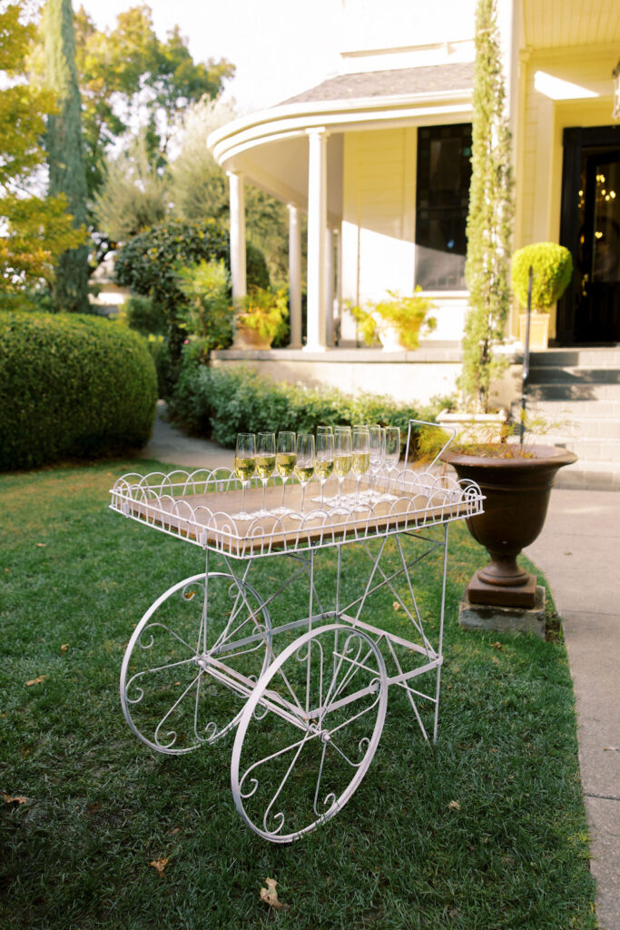 A white metal cart with wheels holds several empty wine glasses and a bowl, set on a lawn near a yellow house with a porch and greenery in the background.