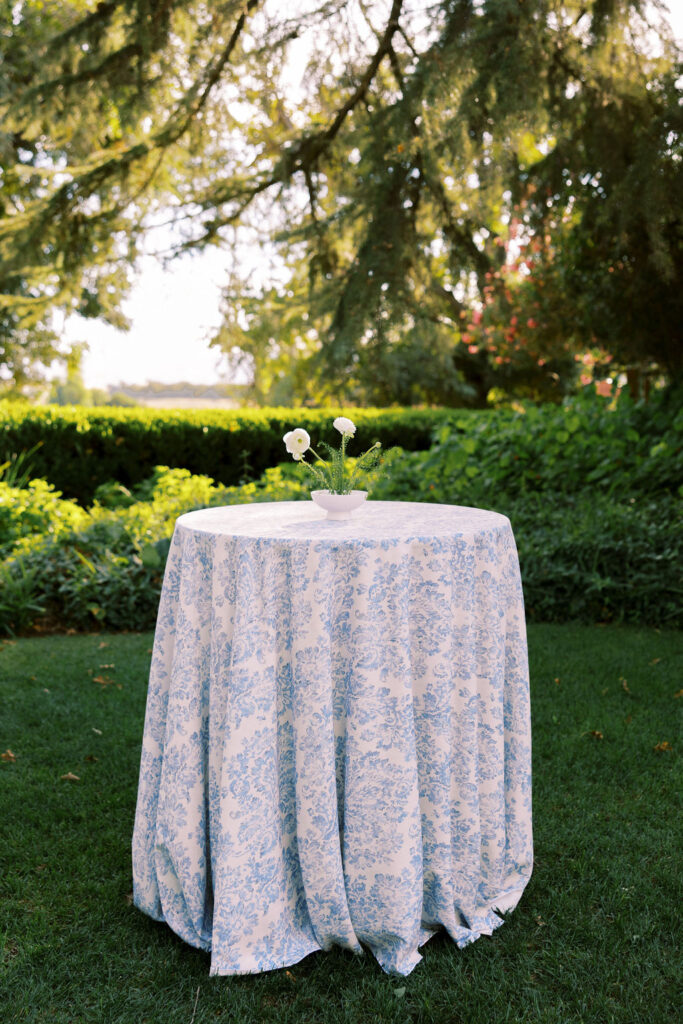 A small round table with a blue and white patterned tablecloth stands outdoors on grass, holding a small vase with white flowers and greenery. Lush trees and bushes are in the background.