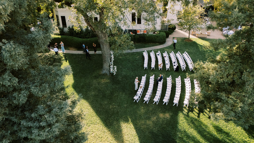 Aerial view of an outdoor wedding ceremony setup with white chairs arranged in rows on a lawn, a few people standing and sitting, and trees casting long shadows.