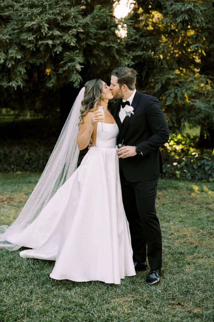 A bride in a white gown and veil kisses a groom in a black suit and bow tie outdoors, both holding drinks, with trees in the background.