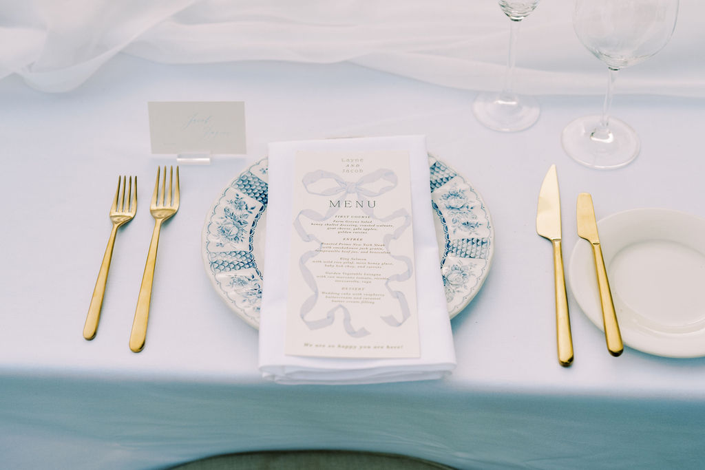 A place setting with a blue and white plate, gold cutlery, a menu on a white napkin, two wine glasses, and a name card on a white tablecloth.