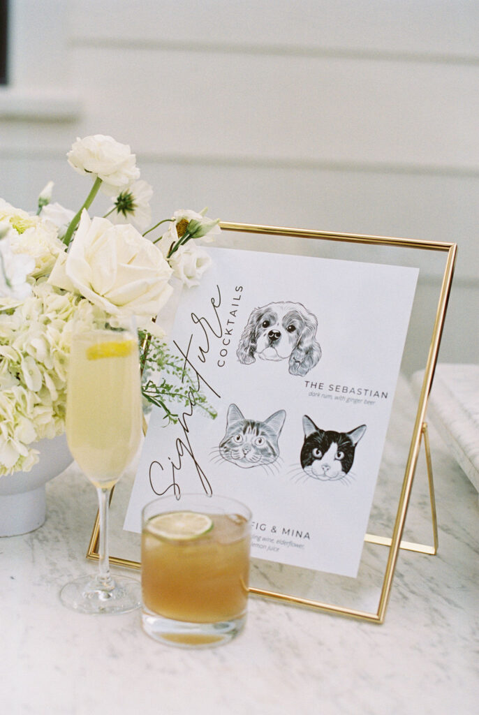 A framed cocktail menu with pet illustrations stands on a marble surface beside a white flower arrangement and two cocktails, one in a flute and one in a rocks glass.