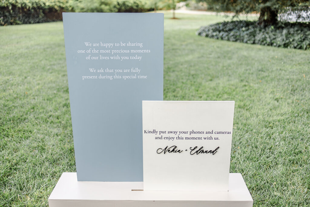 Two wedding signs on display outdoors; one requests guests to be present, and the other asks them to put away phones and cameras and enjoy the moment.