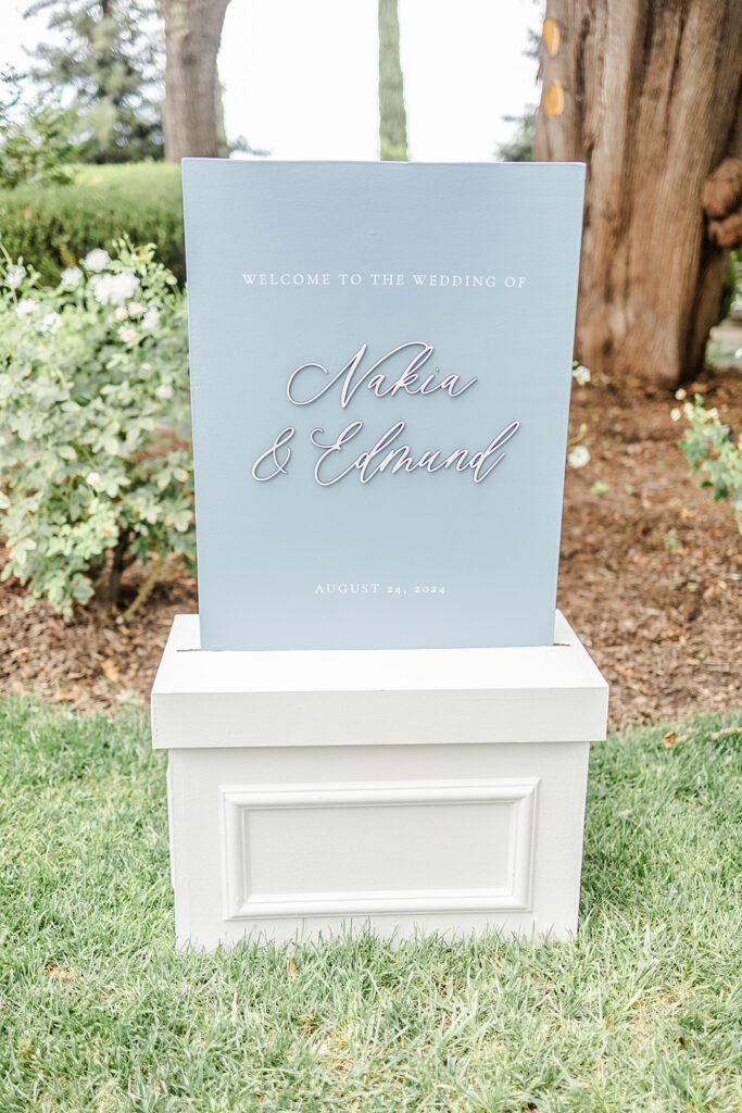 A wedding welcome sign reads "Welcome to the wedding of Nakia & Edmund, August 24, 2024," displayed outdoors on a white stand.