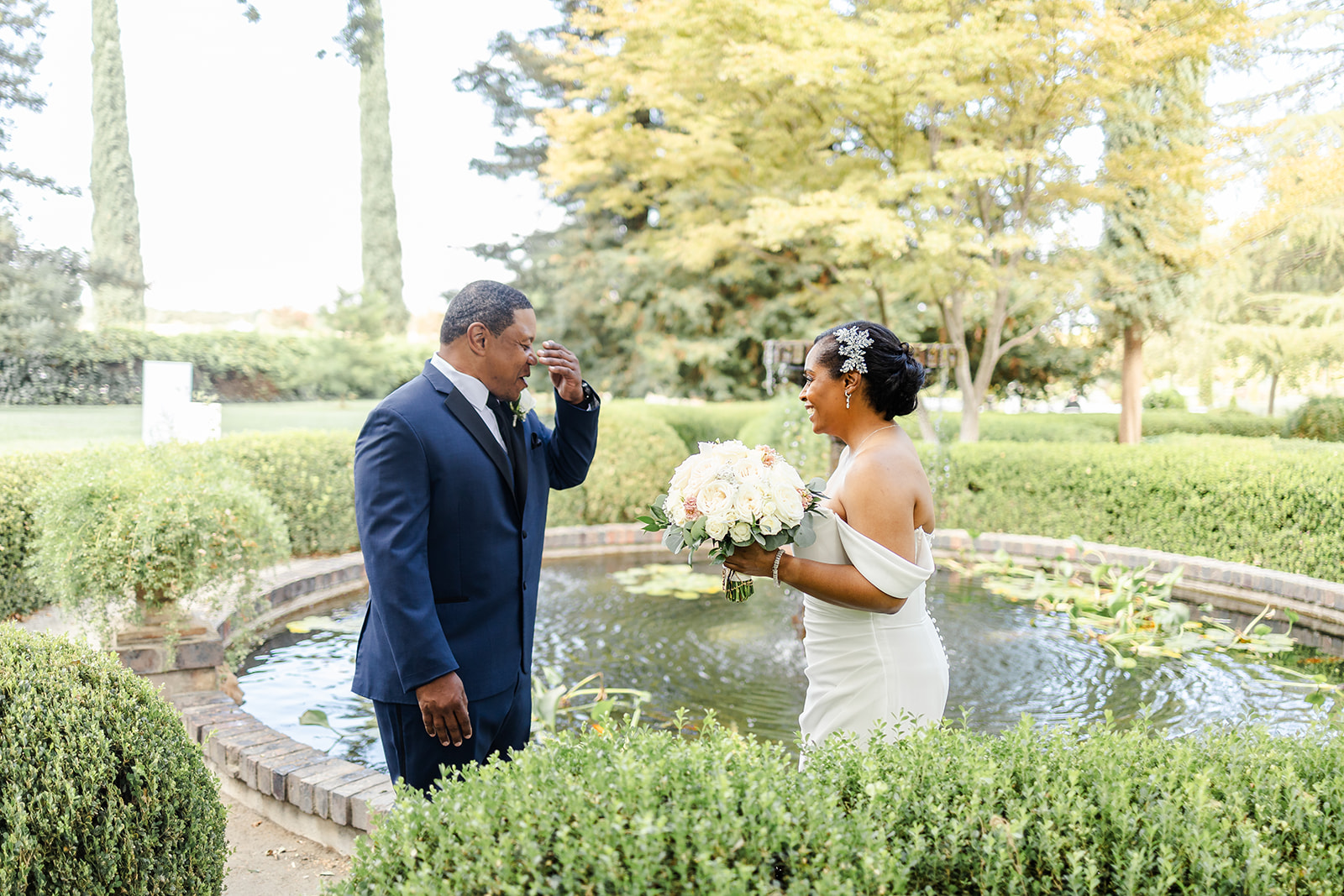 A bride in a white dress holding a bouquet smiles at a groom in a navy suit wiping his eye, standing by a small pond in a garden.