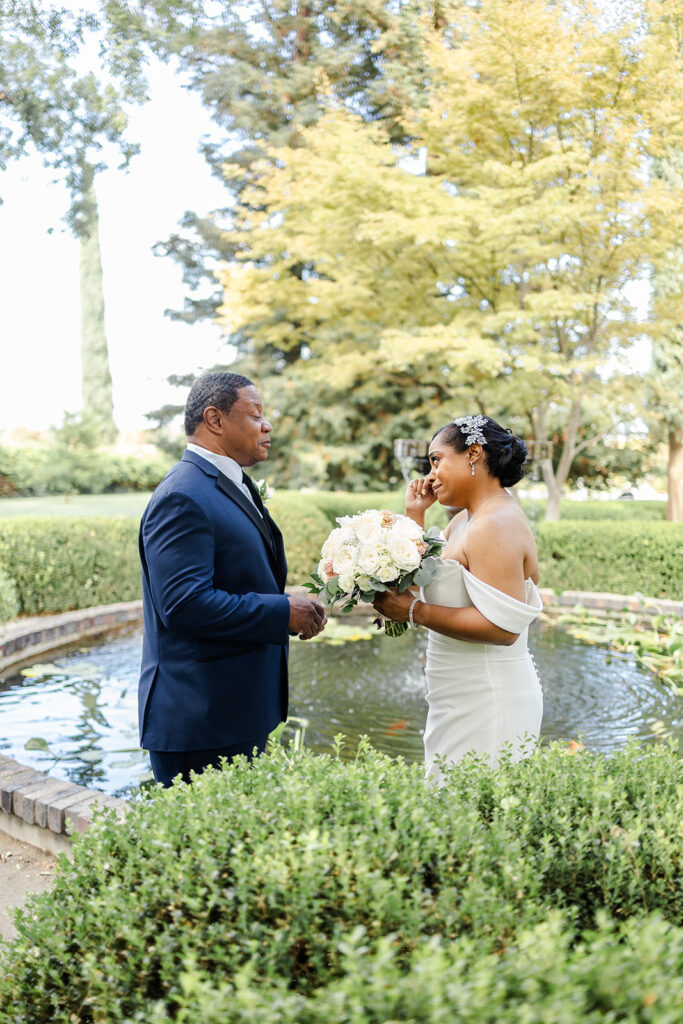 A bride and groom stand by a circular pond, facing each other; the bride holds a bouquet and wipes a tear, while greenery surrounds them.
