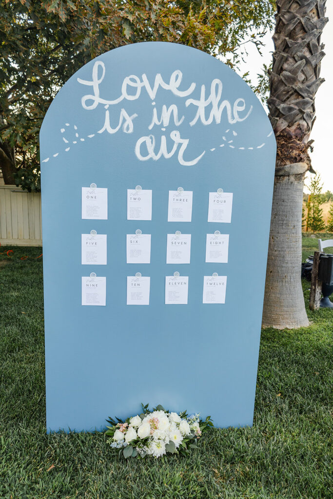 A blue sign with "Love is in the air" written at the top, displaying twelve clipped cards in rows, standing on grass with a white floral arrangement at the base.