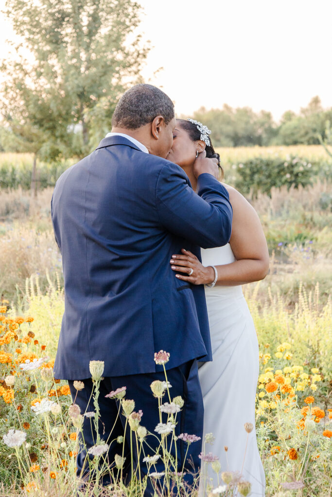 A couple dressed in formal attire shares a kiss in a garden with orange and white flowers, with greenery and trees in the background.