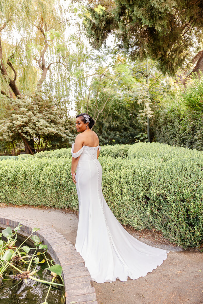A woman in a white off-shoulder wedding dress stands on a garden path near a pond, surrounded by lush greenery and trees.