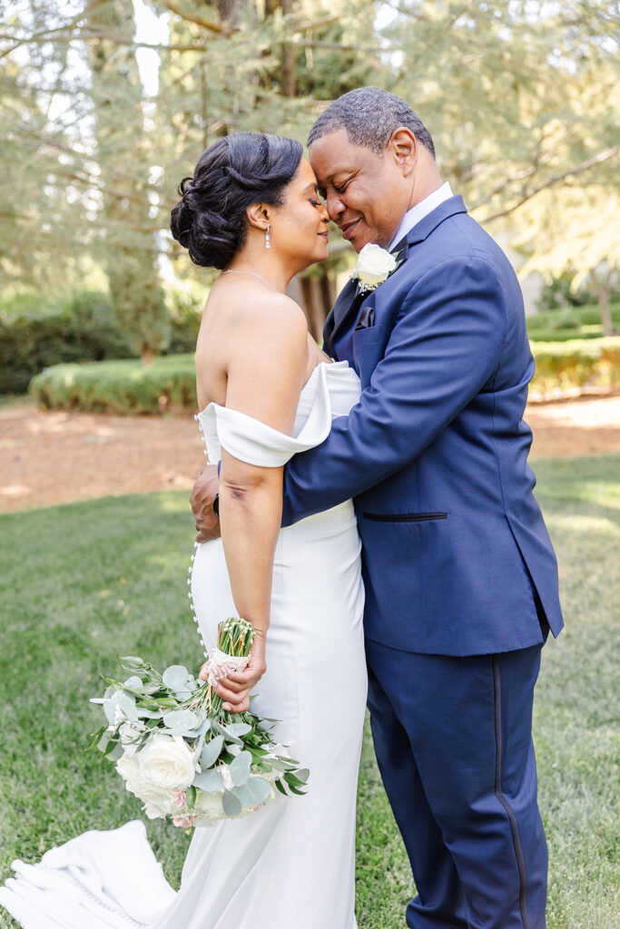 A bride and groom in formal attire embrace outdoors, touching foreheads and smiling, with the bride holding a bouquet of white roses and greenery.