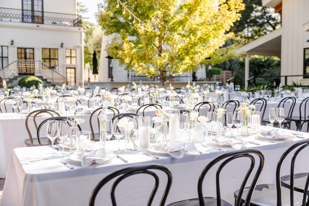 Rows of outdoor tables set with white tablecloths, glassware, candles, and white flowers are arranged in a courtyard with black chairs and greenery in the background.