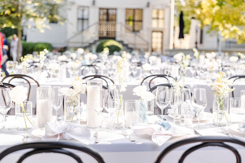 Long tables set for an outdoor event with white tablecloths, glassware, candles, and simple white floral centerpieces; a large building and greenery in the background.