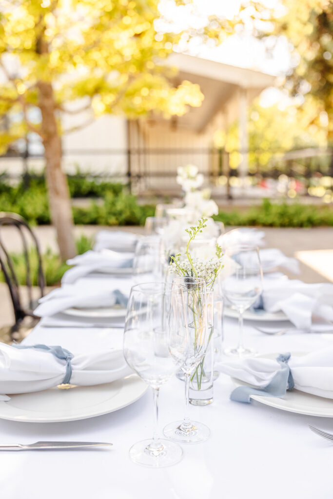 A white tablecloth dining table is set outdoors with plates, napkins, wine glasses, and small vases of white flowers, surrounded by greenery and soft sunlight.