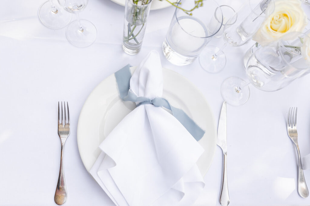A white plate with a neatly folded white napkin and blue ribbon is set on a white tablecloth, surrounded by silver cutlery, glassware, and a small vase with flowers.