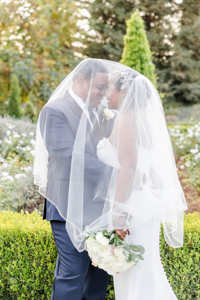 A bride and groom stand in a garden, facing each other and smiling under a shared veil. The bride holds a bouquet of white flowers.