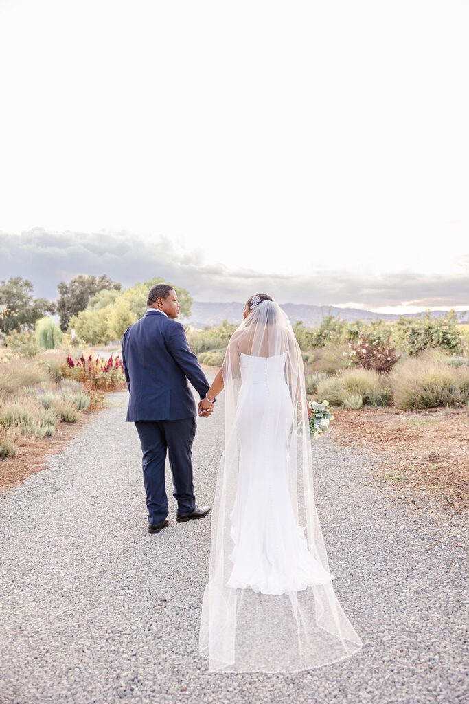 A bride in a white dress and veil holds hands with a groom in a navy suit as they walk down a gravel path in a garden setting.