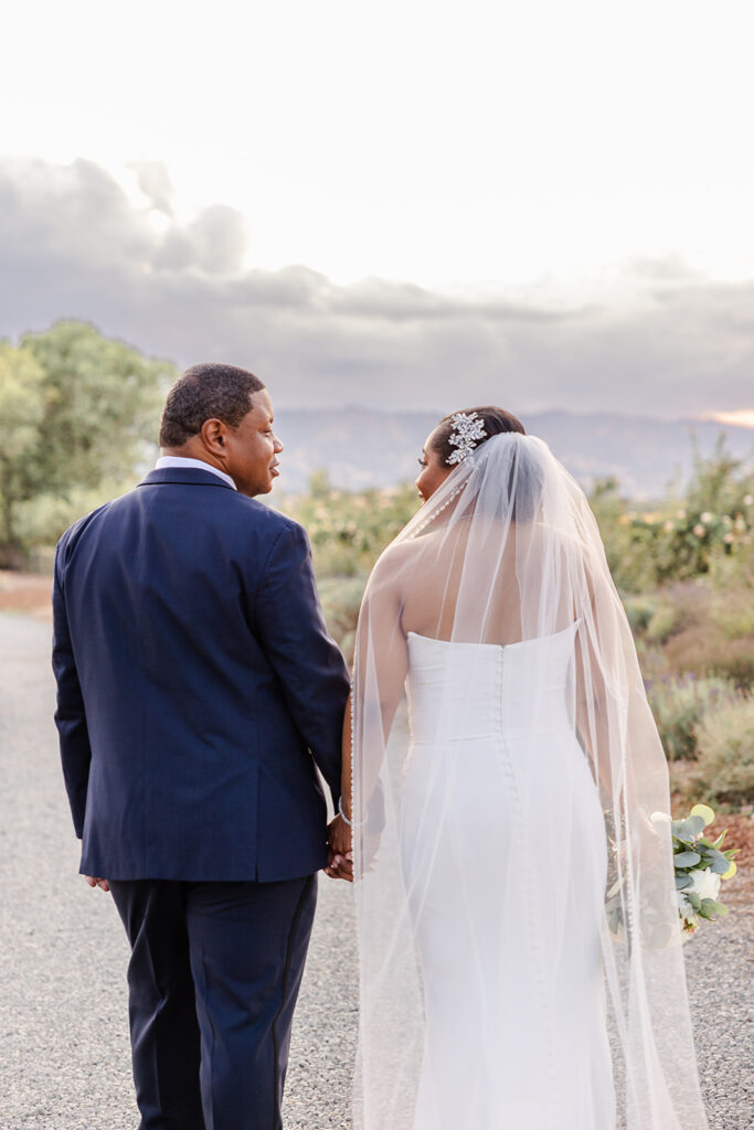 A bride and groom walk hand in hand outdoors on a path, facing away from the camera. The bride wears a white dress and veil, while the groom wears a dark suit.