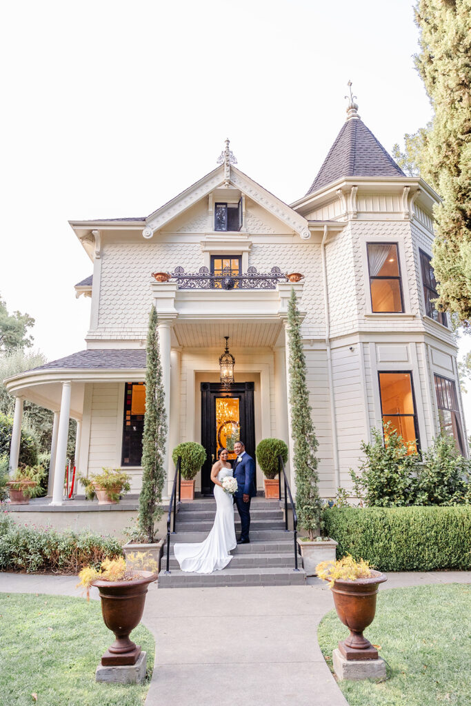 A couple in wedding attire stands on the front steps of a large, light-colored Victorian-style house surrounded by greenery.