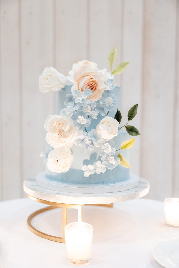 A light blue tiered cake decorated with white and peach sugar flowers and green leaves sits on a marble stand, surrounded by small lit candles.