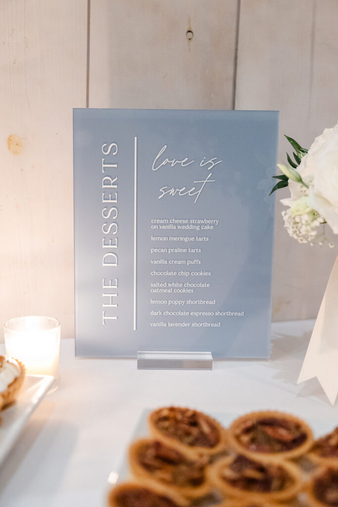 A dessert menu sign lists various sweets such as cream cheese strawberry, lemon meringue tarts, and chocolate chip cookies on a table with candles, flowers, and pecan tarts.
