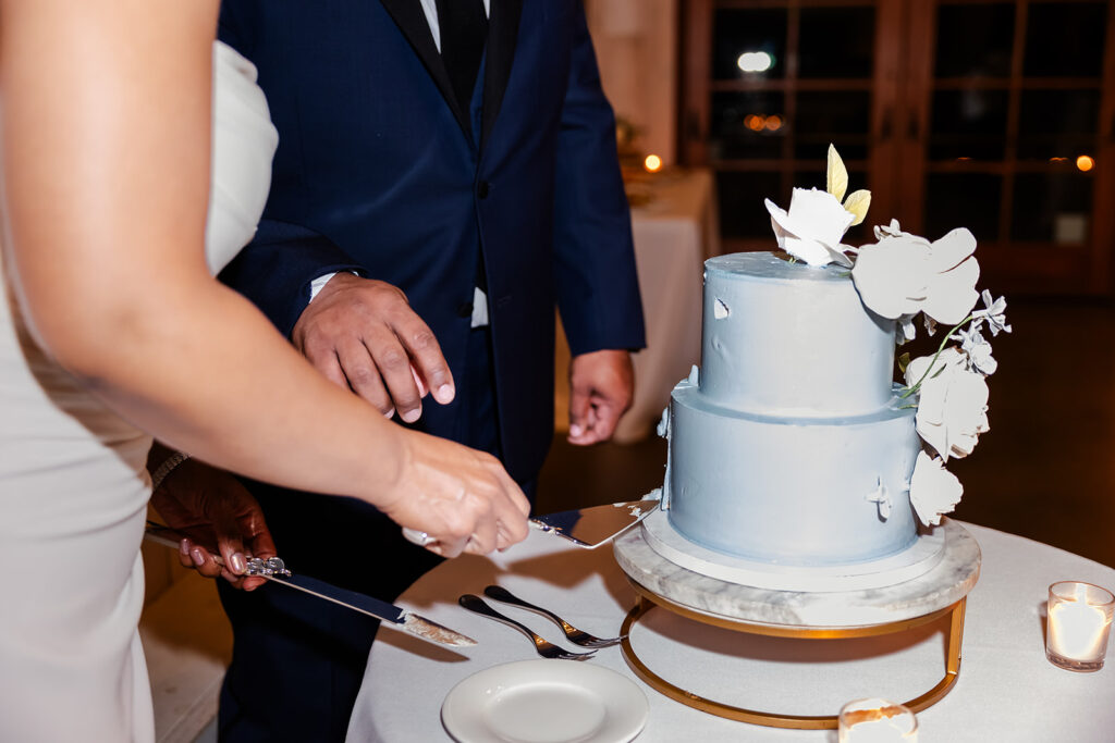 Two people, dressed formally, are cutting a two-tiered light blue cake with white floral decorations at an event.