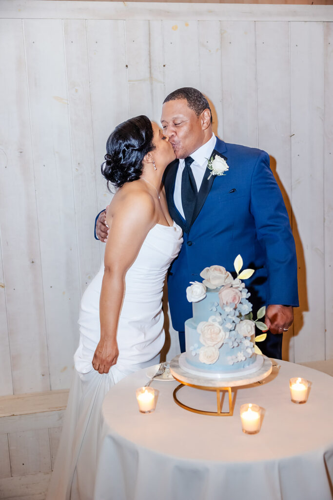 A couple dressed in wedding attire kisses beside a decorated cake on a table with lit candles, in front of a white wooden wall.