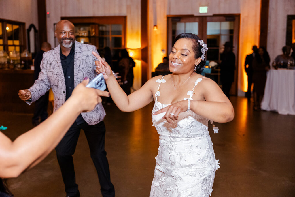 A woman in a white dress dances and smiles at an indoor event, with other people visible in the background.