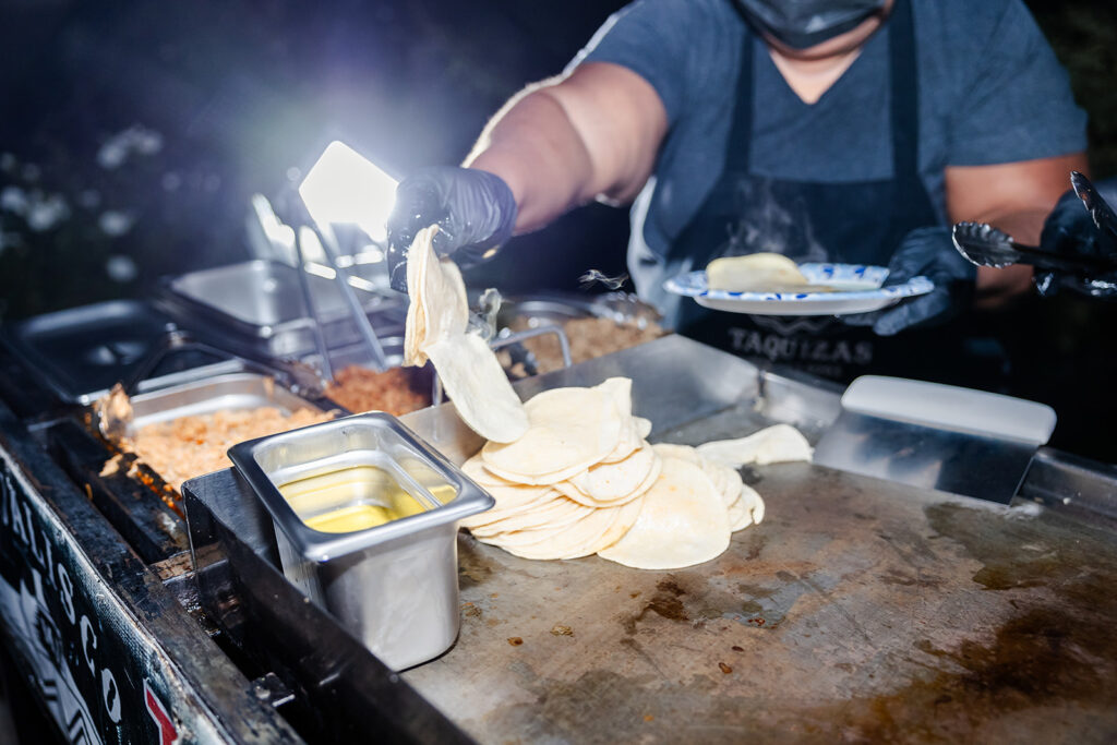 A person wearing gloves and a mask serves tortillas from a grill at a food stand, with various food trays and a plate visible.