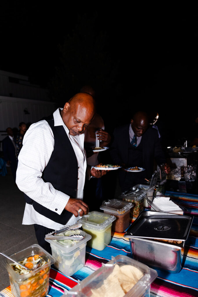 A man in a white shirt and black vest serves himself food from a buffet table outdoors at night.