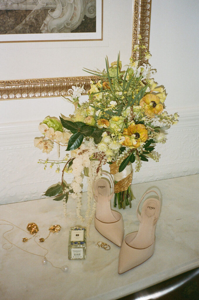 A bouquet of yellow and white flowers, nude Fendi heels, rings, earrings, a necklace, and a perfume bottle arranged on a marble surface below a framed picture.