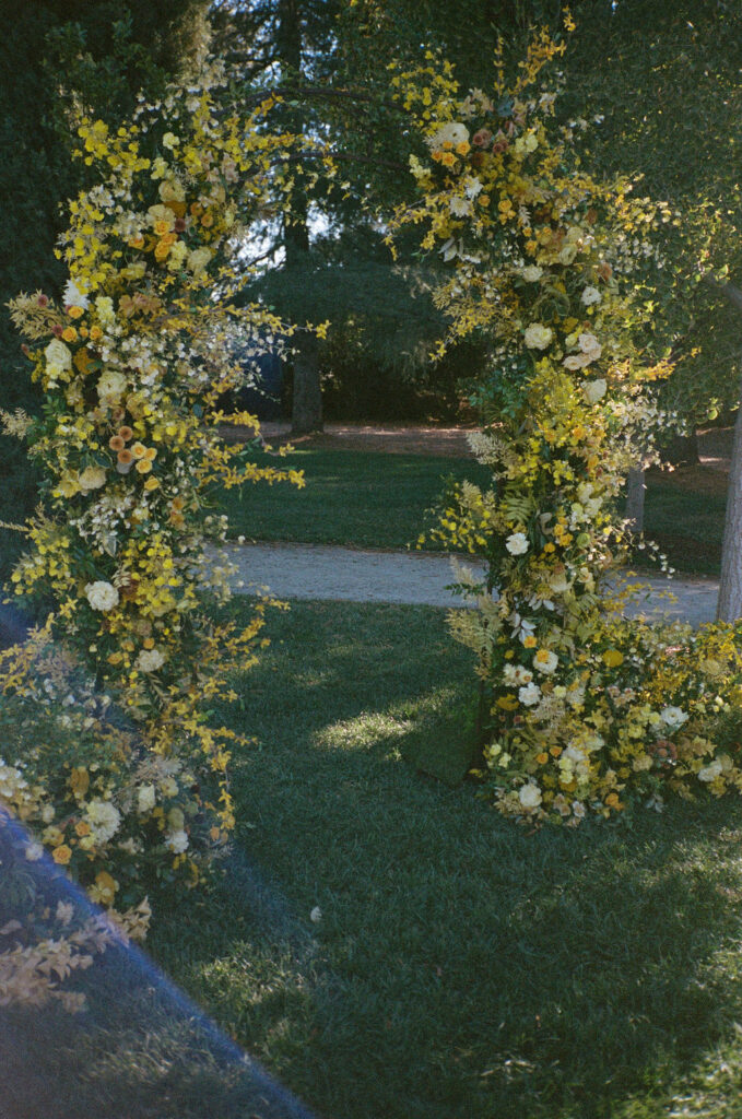 Two tall floral arches decorated with yellow and white flowers stand on a grassy lawn with trees and a path in the background.