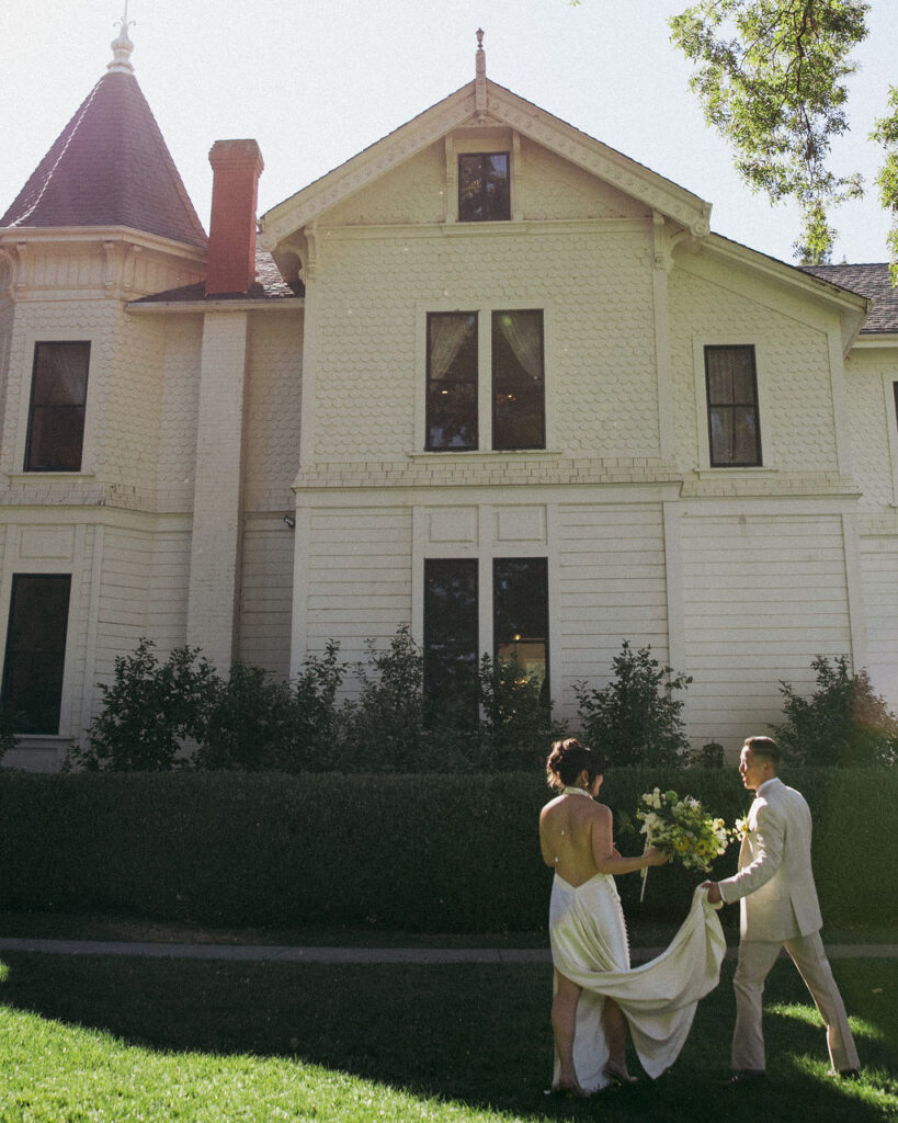 A couple dressed in formal attire walks on the lawn in front of a large, white Victorian-style house on a sunny day.