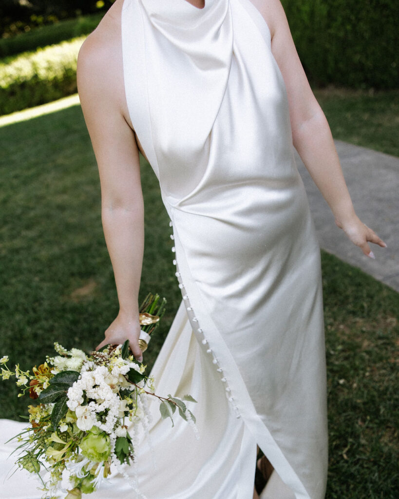 A person in a sleeveless white satin dress with side buttons stands outdoors on grass, holding a bouquet of flowers in one hand.