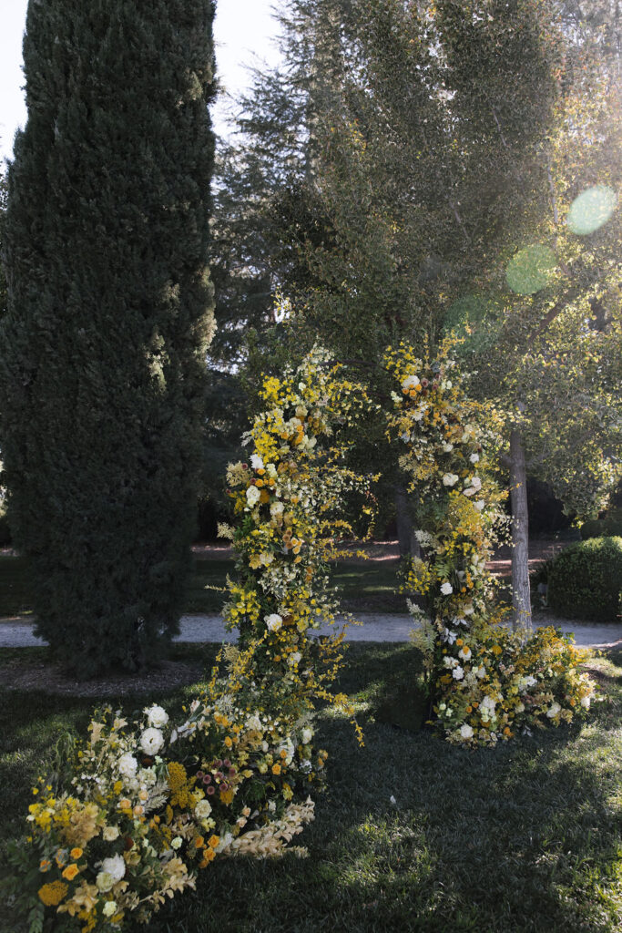 Tall floral arch made of yellow and white flowers stands on grass in a sunlit garden, framed by large trees and greenery.