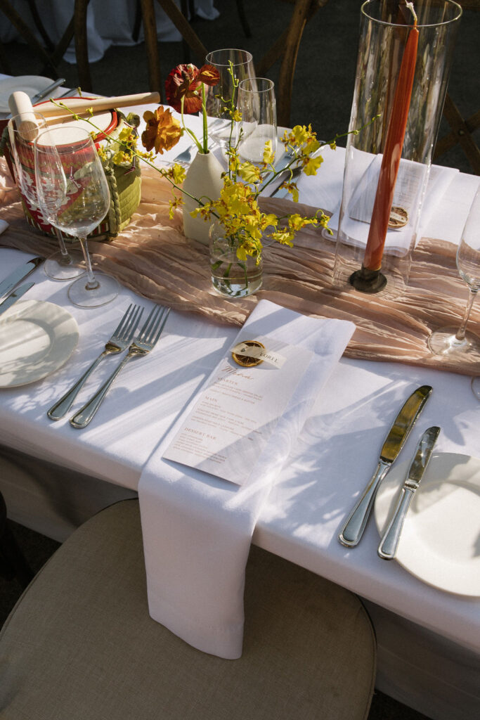 Elegant table setting with a white tablecloth, gold cutlery, wine glasses, a menu on a napkin, floral centerpieces, and tall candles in glass holders.