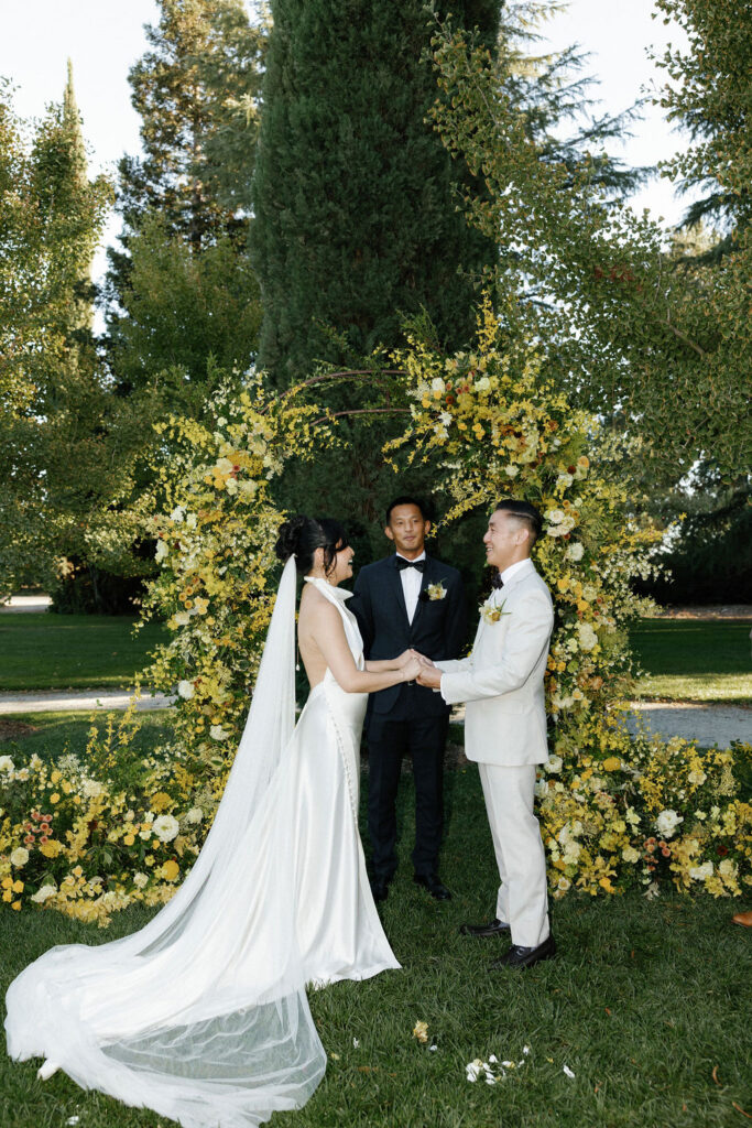 A bride and groom stand holding hands during their wedding ceremony outdoors, with an officiant and a floral arch behind them.