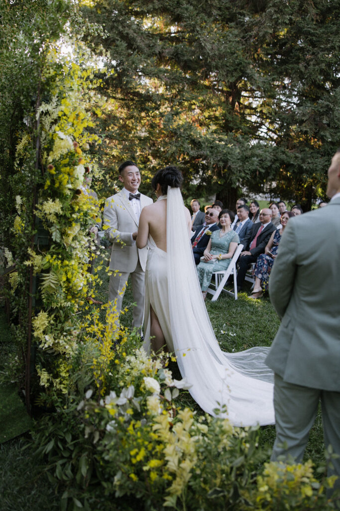 A bride and groom stand facing each other during an outdoor wedding ceremony, surrounded by flowers and seated guests.