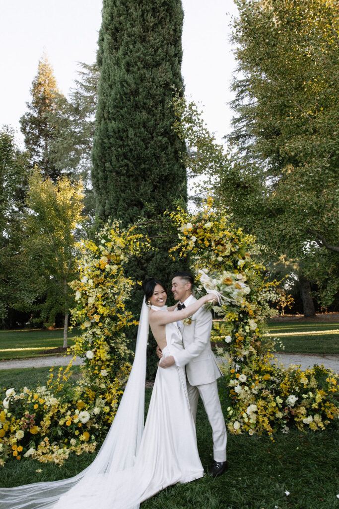 A bride and groom embrace and smile in front of a floral arch outdoors, surrounded by yellow flowers and greenery.