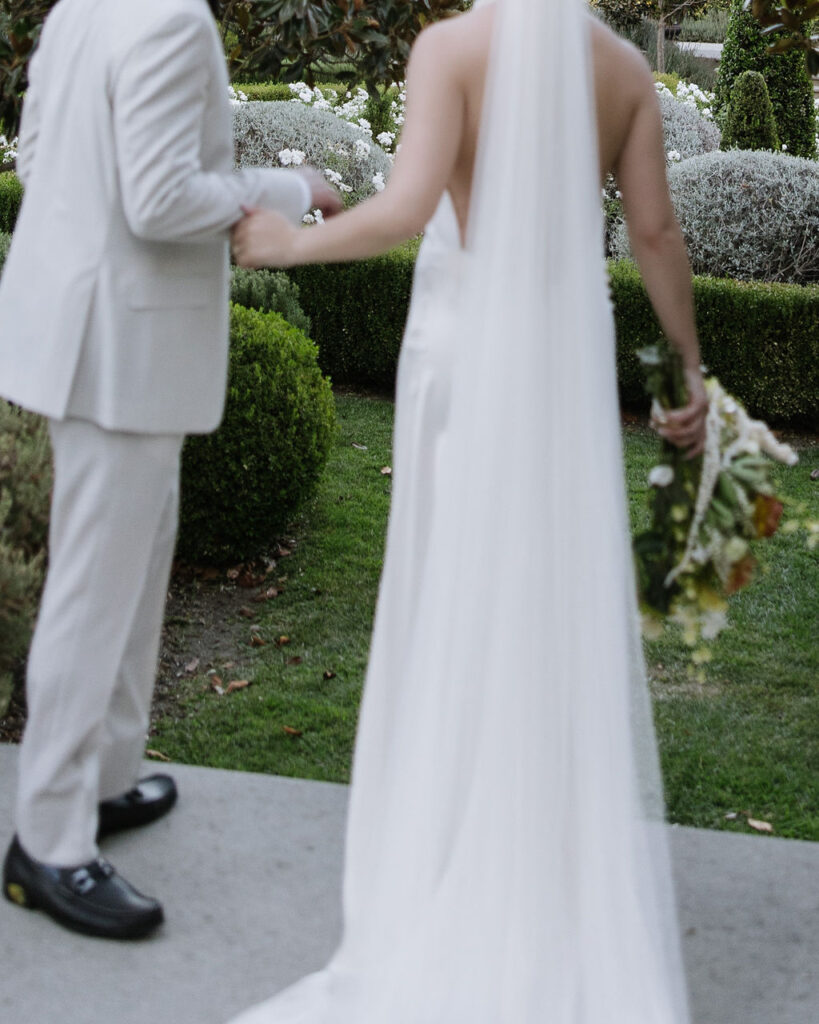 A bride and groom stand outdoors holding hands. The groom wears a light suit; the bride wears a backless gown and veil, holding a bouquet. Their faces are out of focus.