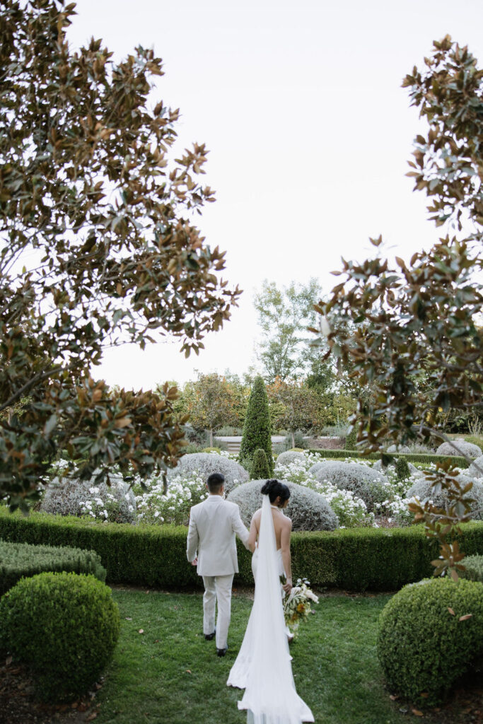 A bride and groom walk hand in hand through a manicured garden, surrounded by greenery and flowers, with the bride holding a bouquet and wearing a long white veil.