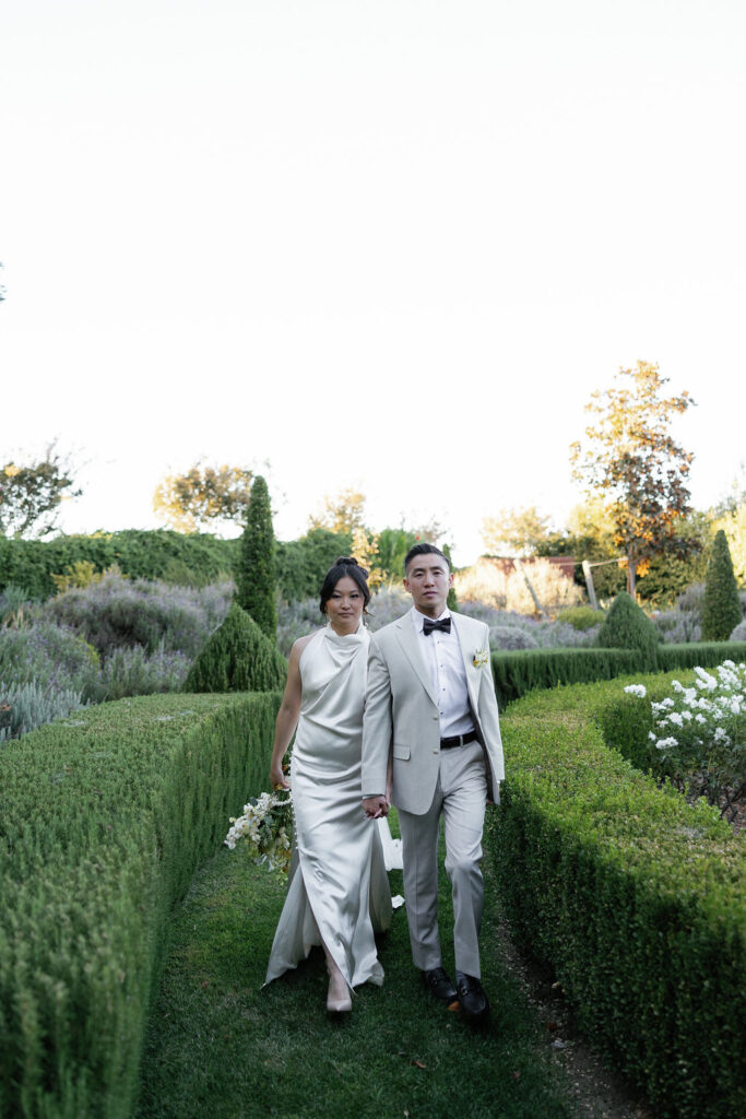 A couple in formal attire walks through a manicured garden, holding hands. The woman wears a white dress and holds flowers; the man wears a light suit with a black bow tie.