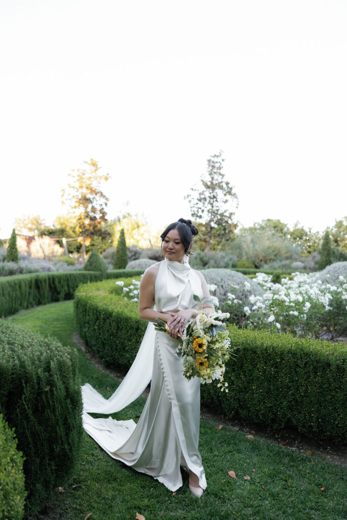 A woman in a white wedding dress holds a bouquet of flowers while standing on a garden path lined with green hedges and white flowers.