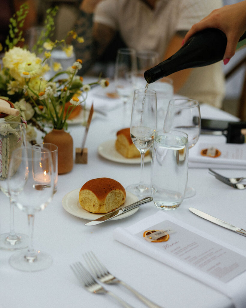 A hand pours sparkling wine into a glass on a white-tablecloth dining table set with bread rolls, menus, flowers, candles, and glassware.