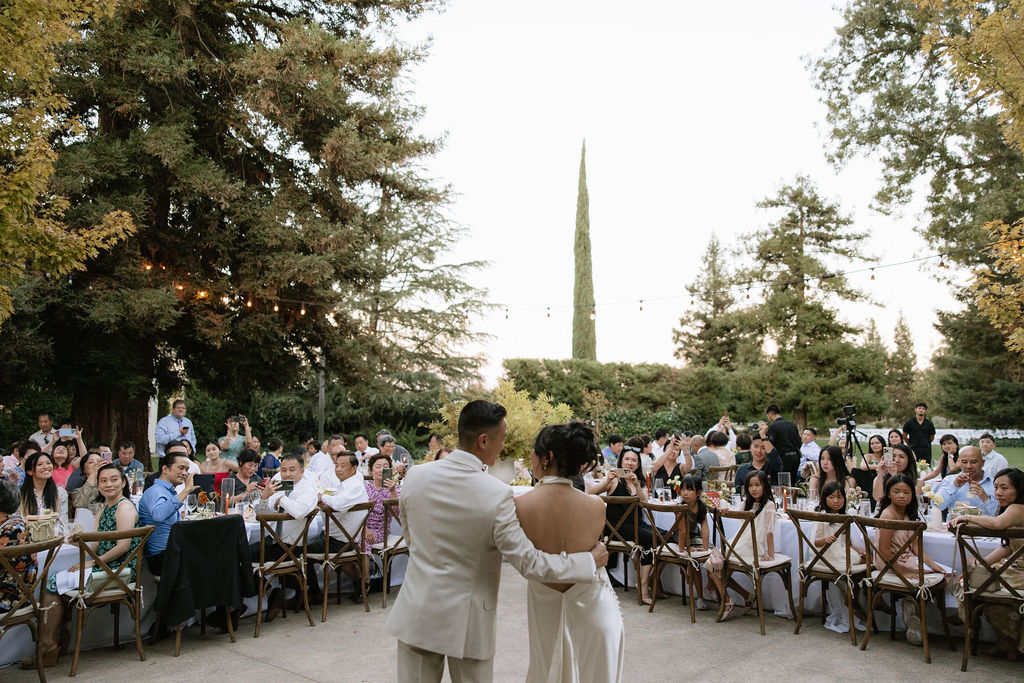 A couple stands together facing seated guests at an outdoor event, surrounded by trees and string lights, during daylight.