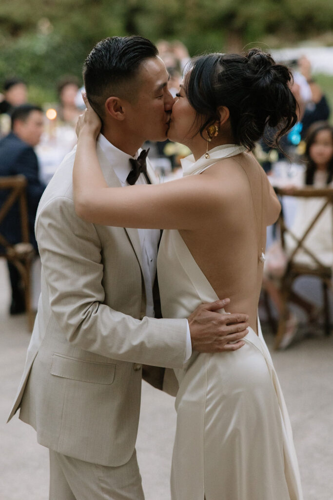 A couple dressed formally shares a kiss while dancing at an outdoor event, with seated guests visible in the background.